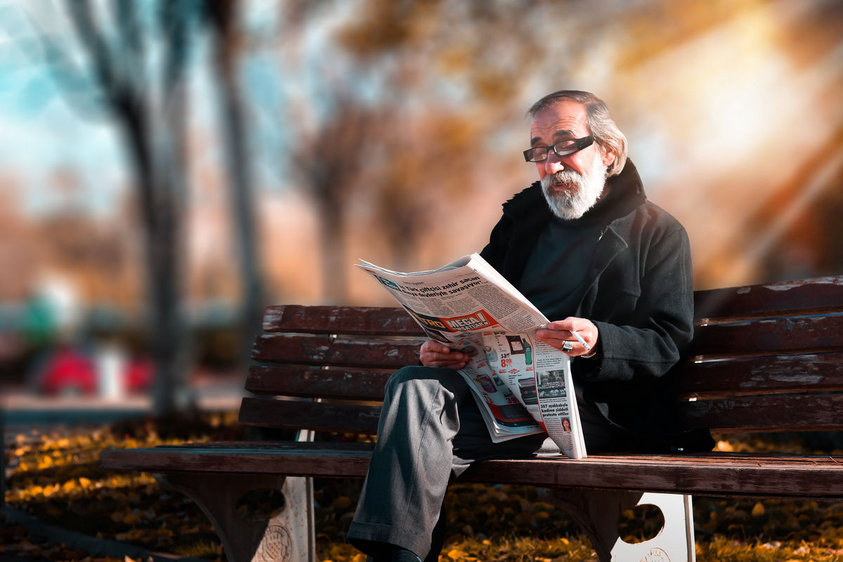 Relaxed older man with a beard on a park bench reading newspaper.