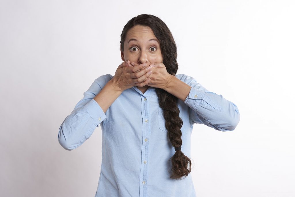 Privacy - Say NOTHING! Woman in blue long sleeve shirt with long braided hair shocked and covering her mouth with both hands.