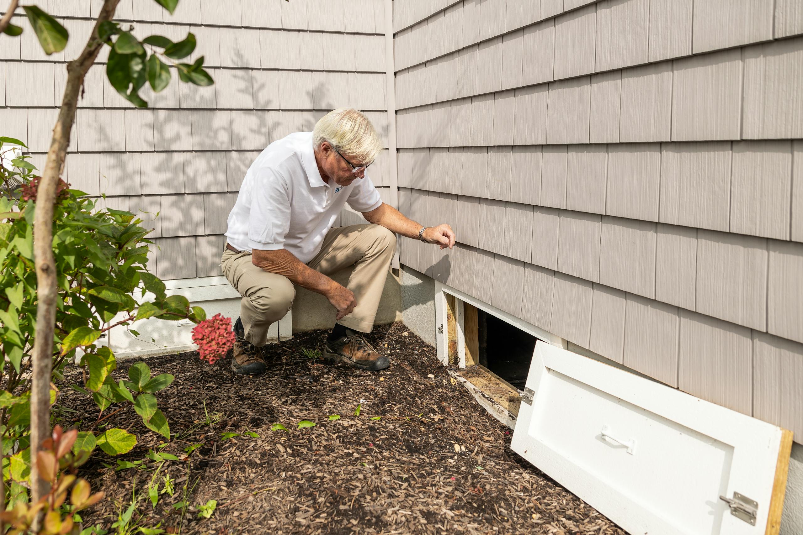 Senior adult inspecting a crawl space entrance during a home inspection on a sunny day.