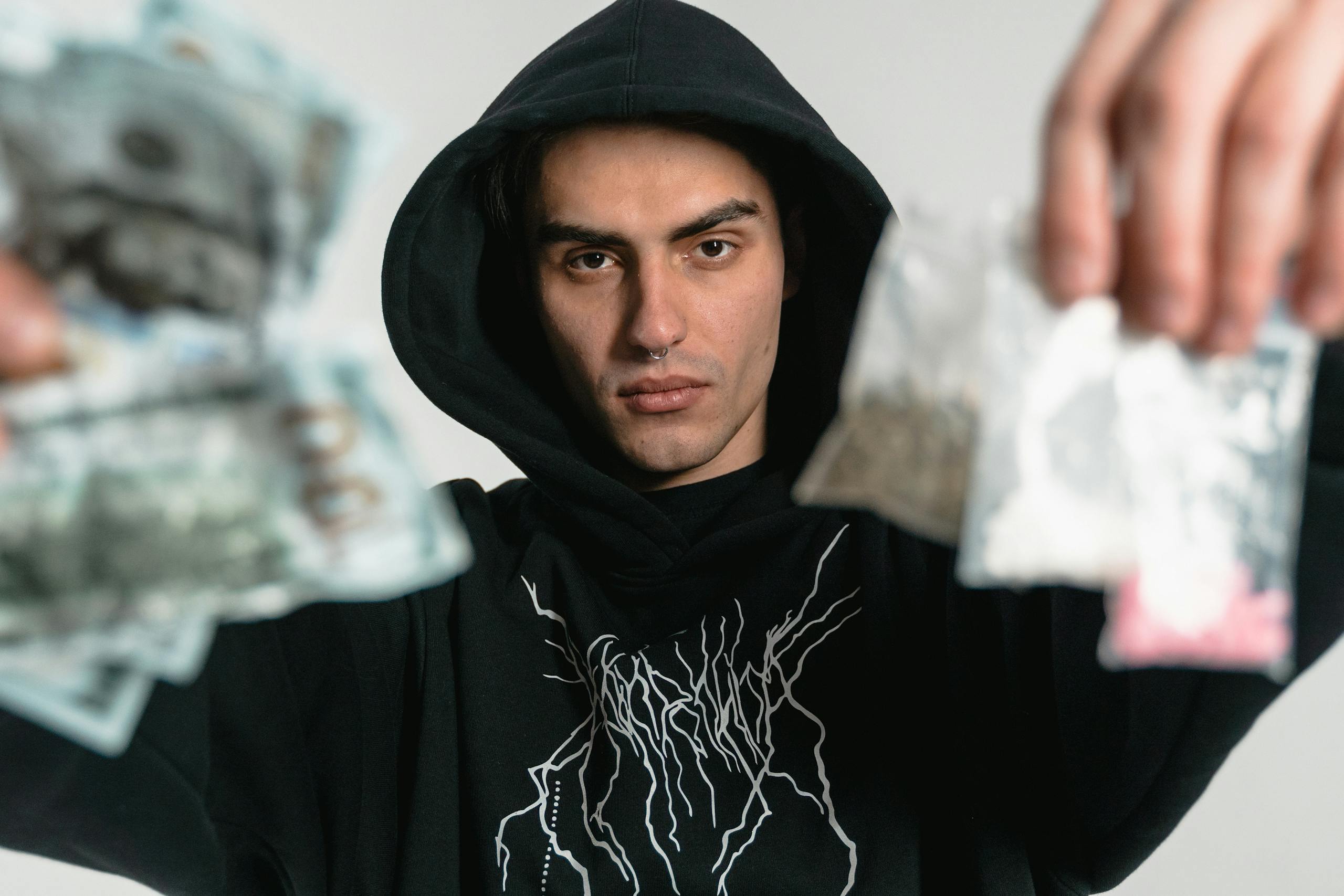 Portrait of a man in a black hoodie holding cash and bags of drugs, depicting illegal trade